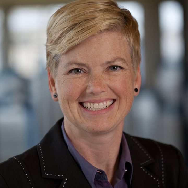 A person with short blonde hair, wearing a black blazer and purple shirt, smiles at the camera in a blurred indoor setting.