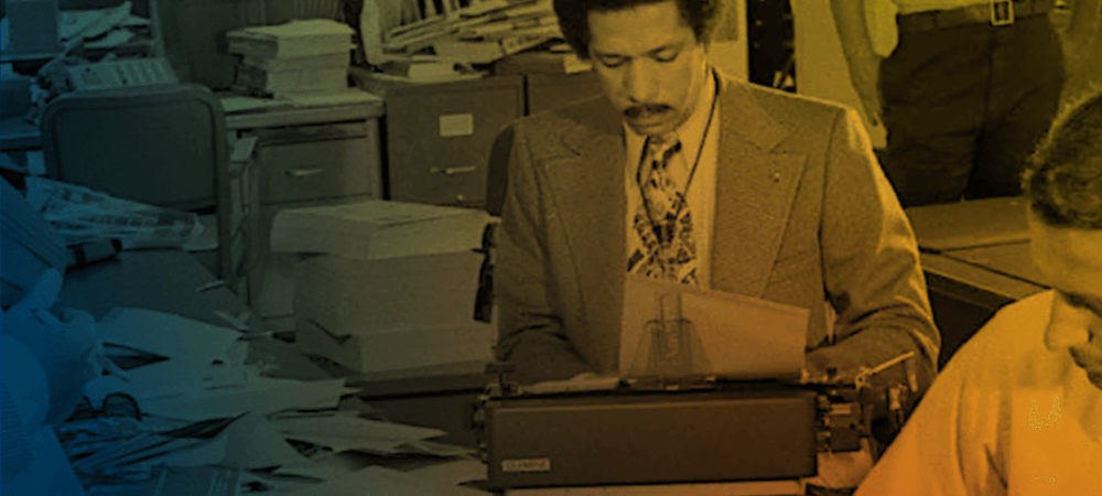 Two men work at cluttered desks filled with papers in a busy office. One man types on a typewriter while another stands in the background with arms crossed.