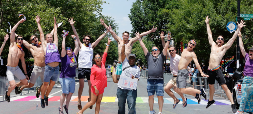 A group of people jump in the air on a rainbow-painted crosswalk during a sunny day, celebrating lgbtqi health care new rules while some hold pride flags and wear festive attire.