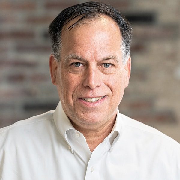 A middle-aged man with short dark hair wearing a white button-down shirt, smiling at the camera in front of a blurred brick background.