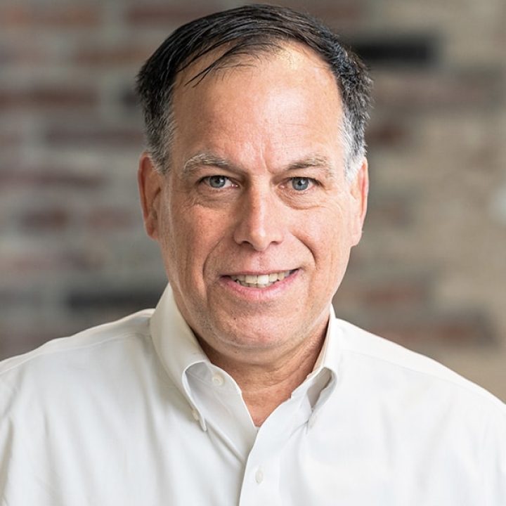 A middle-aged man with short dark hair wearing a white button-down shirt, smiling at the camera in front of a blurred brick background.