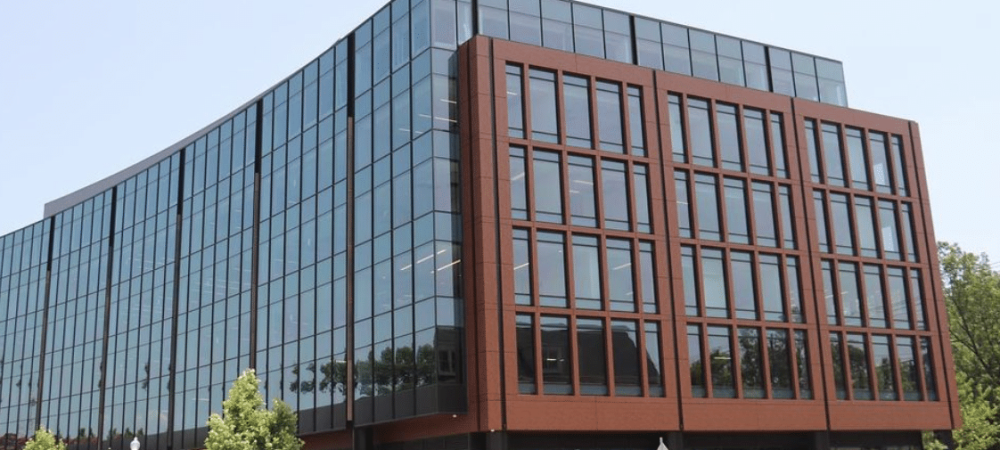 A modern, multi-story office building with large glass windows and a brown frame, located on a city street corner with trees and a few pedestrians nearby.