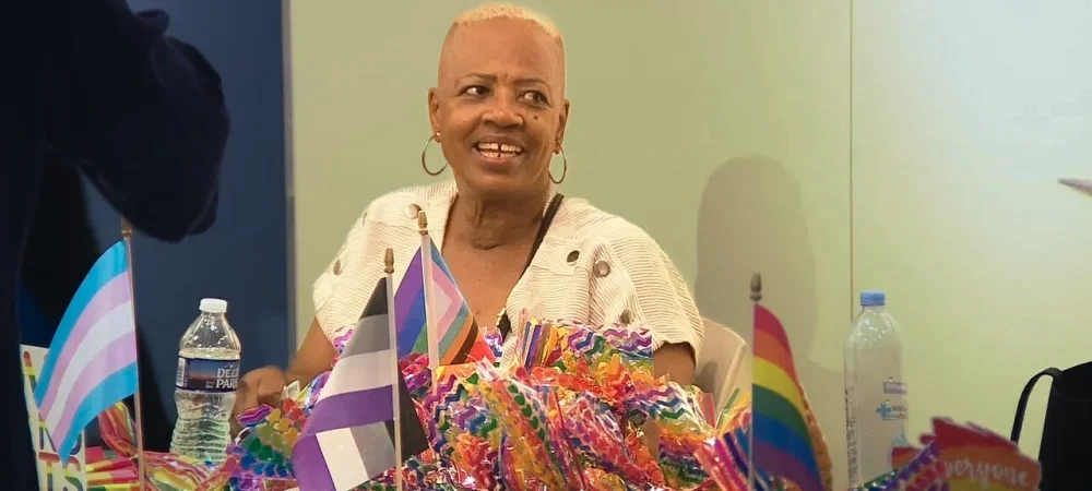 A person with short blond hair sits at a table covered with various pride flags and colorful decorations, smiling, with a water bottle and brochures nearby.