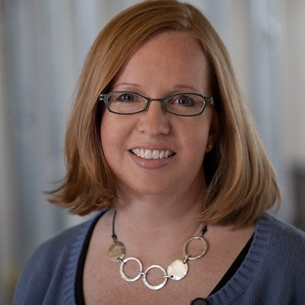 A woman with shoulder-length red hair, glasses, and a blue sweater smiles at the camera. She is wearing a necklace with large circular links. The background is softly blurred.