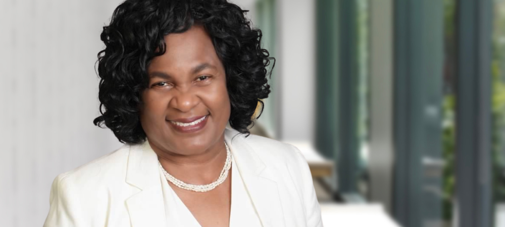 A woman with curly black hair wearing a white blazer, white top, and pearl necklace stands in a bright office hallway.