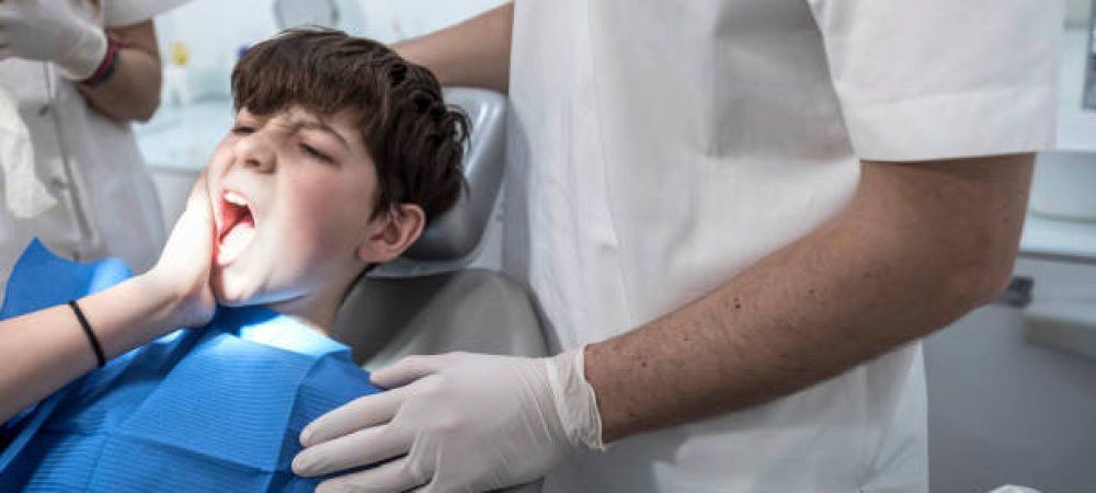 A boy sits in a dental chair holding his cheek in pain while a dentist in white gloves stands beside him.