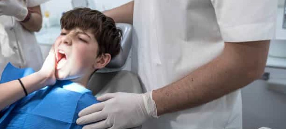 A boy sits in a dental chair holding his cheek in pain while a dentist in white gloves stands beside him.