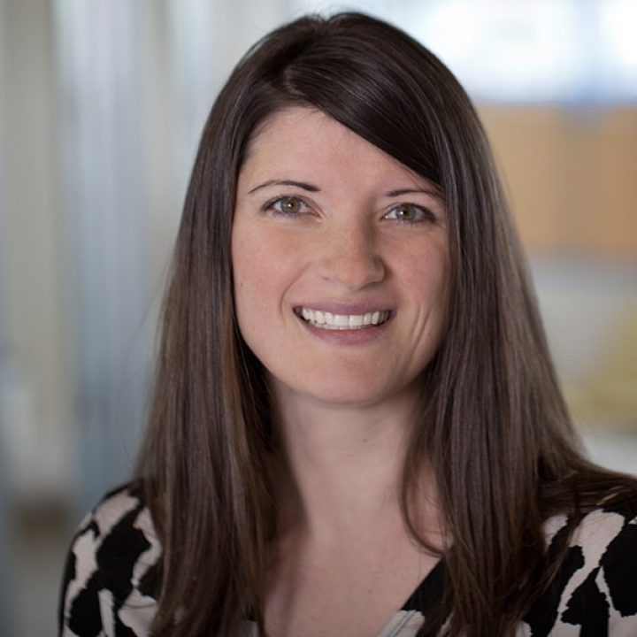 A woman with straight brown hair and a patterned top smiles at the camera in an indoor setting with a blurred background.