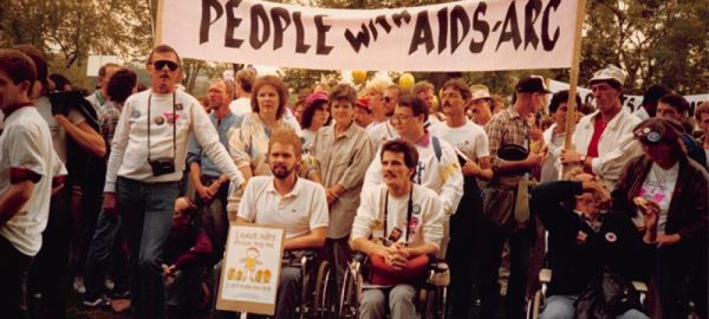 A group of people, including individuals in wheelchairs, hold a banner reading "People with AIDS-ARC" at an outdoor gathering or protest.