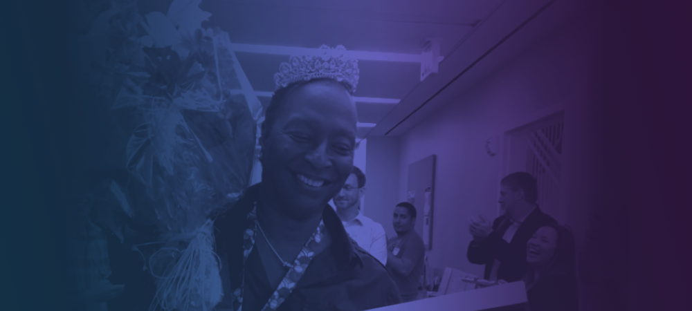 A person wearing a tiara smiles while holding a bouquet, surrounded by people clapping in a hallway.