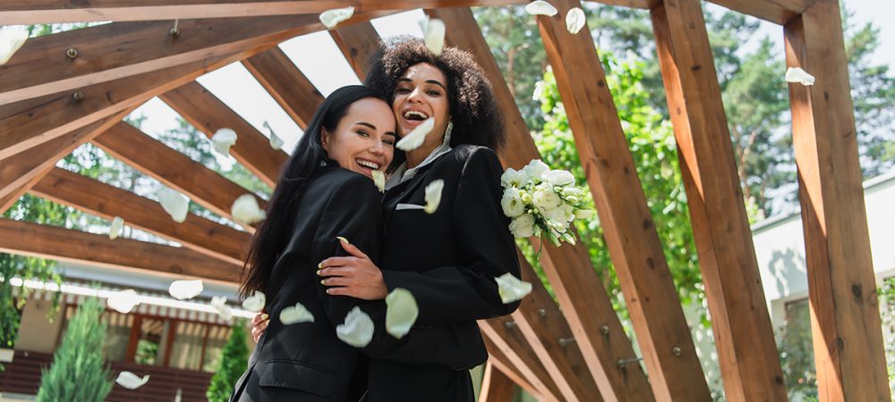 Cheerful multiethnic lesbian couple in suits hugging near petals during wedding in park.