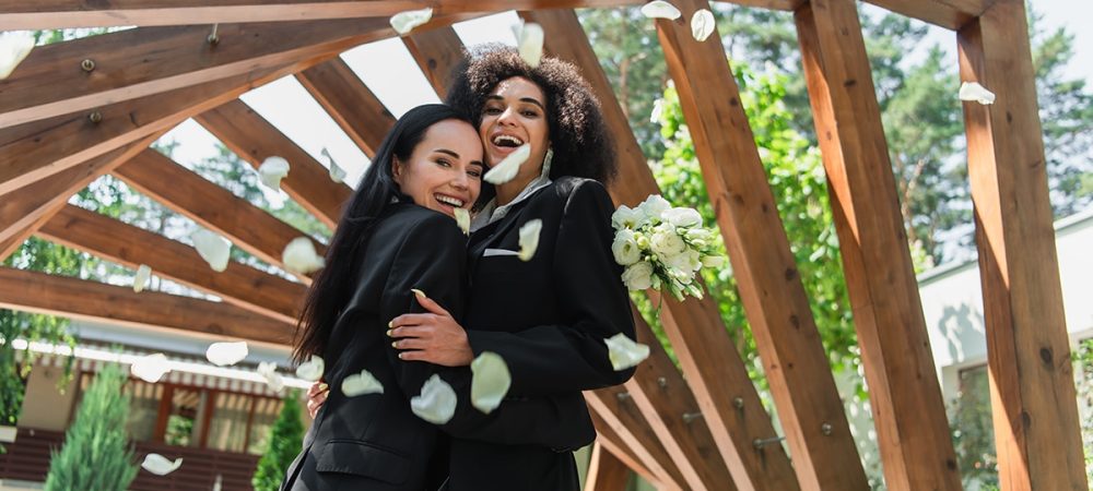Two women in suits hugging under a wooden structure.