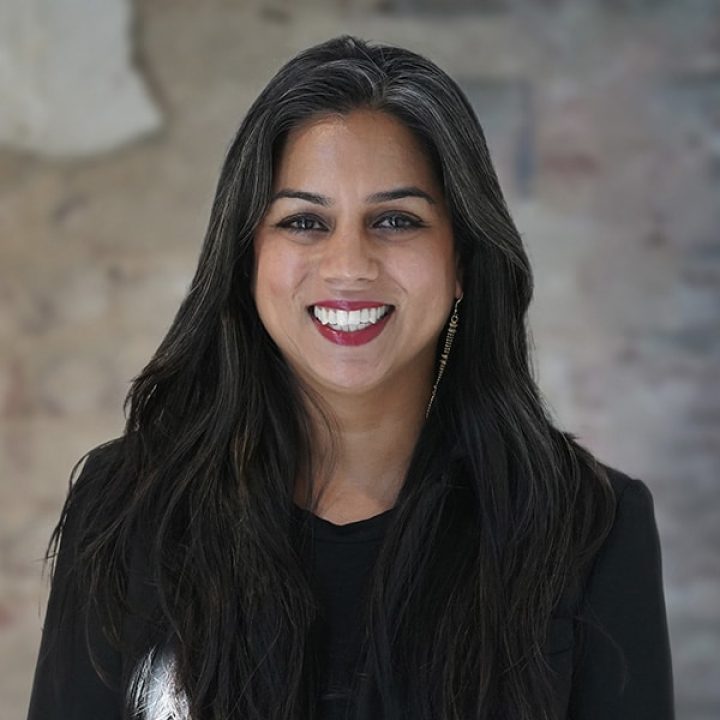 A woman with long dark hair, wearing a black top and blazer, smiles at the camera against a blurred, neutral-toned background.