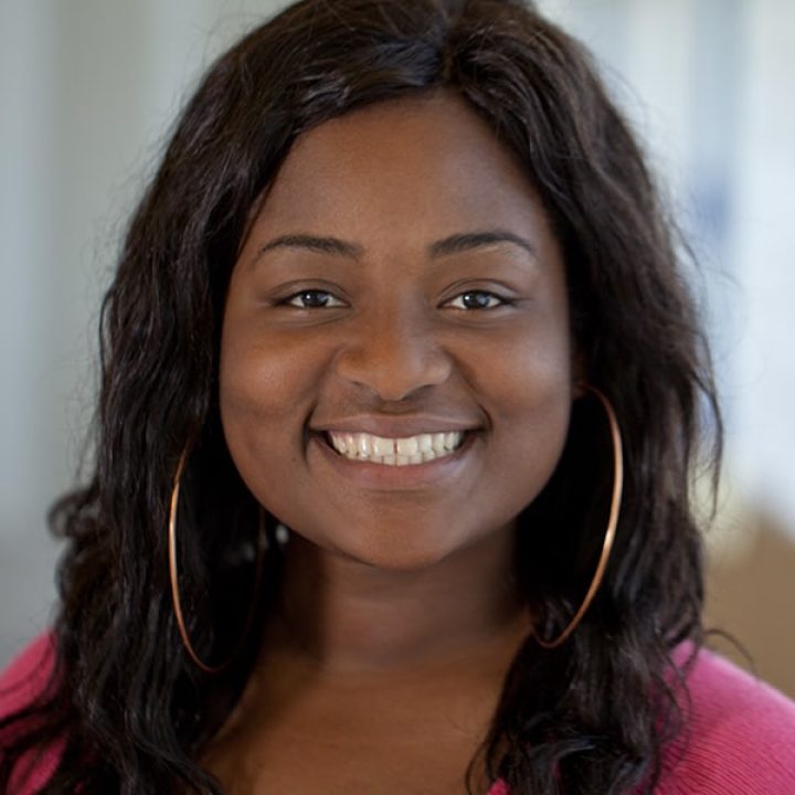 A woman with long dark hair, wearing large hoop earrings and a pink top, smiles at the camera against a blurred indoor background.
