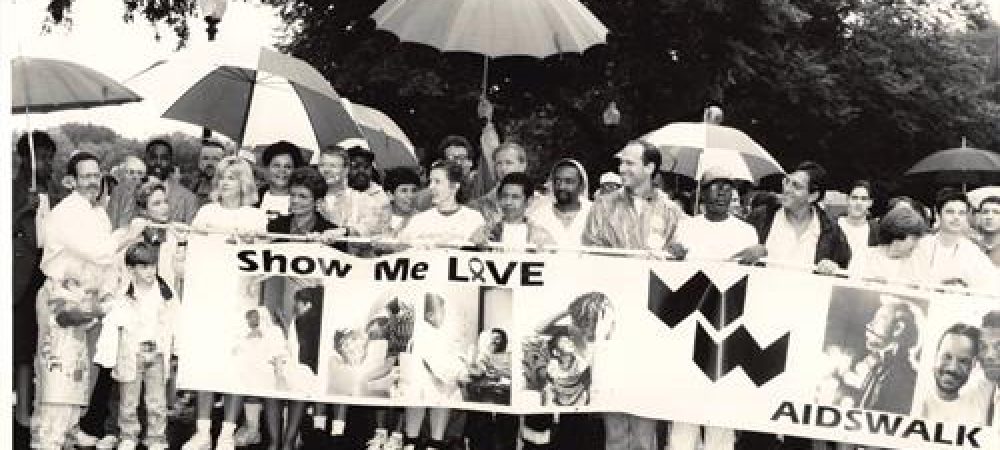 A diverse group of people stand together holding umbrellas and a large "Show Me Love AIDSWALK" banner during a rainy outdoor event.