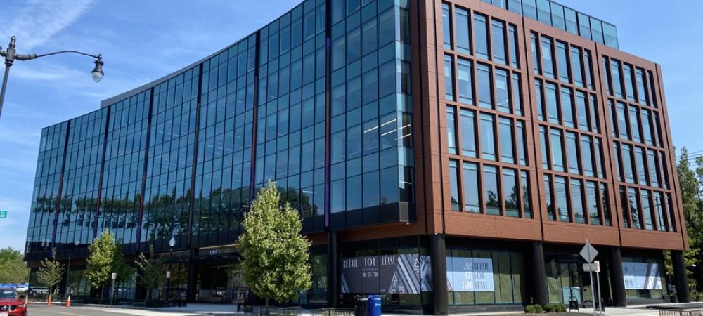 Modern glass and steel office building on a street corner with trees, wide sidewalks, and blue sky in the background.