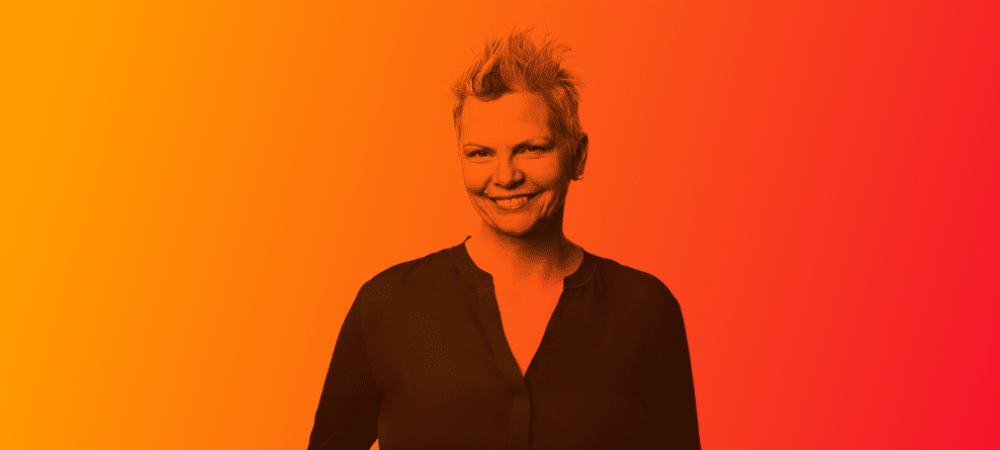 Smiling person with short hair wearing a dark shirt stands against a gradient background transitioning from yellow to red.