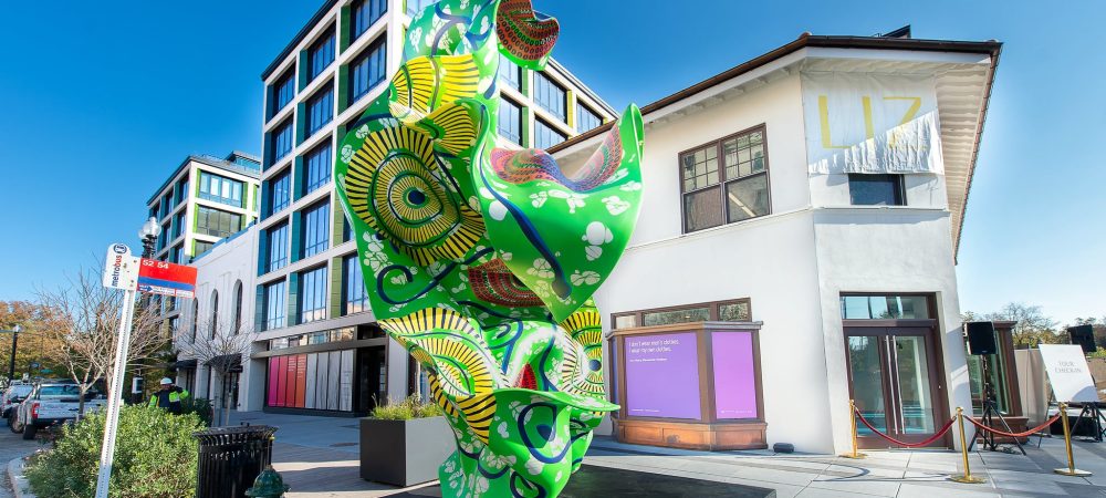 A vibrant, abstract green and yellow sculpture stands on a pedestal in an urban plaza, surrounded by modern and older buildings under a clear blue sky.